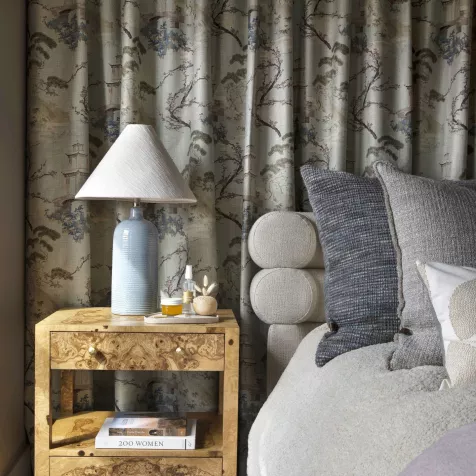 A section of a bedroom with a curtain wall draped behind a bedside table and lamp, which are next to a bed with a fabric headboard and large pillows. 